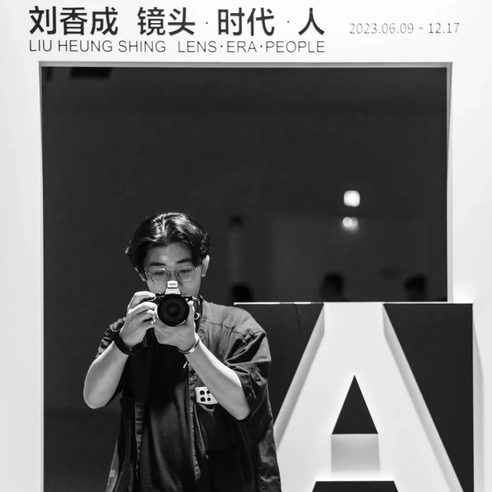 黑白肖像中，一位摄影者举起相机站在展览空间里